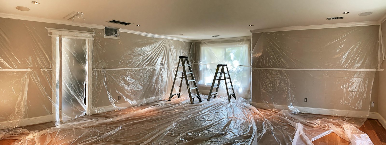 Emptied living room prepared for ceiling work, clear plastic sheeting taped up the walls and laid across the floor, the doorway at one side closed off behind a zippered sheet, a ceiling supply vent taped over and two step ladders standing open under a bare flat white ceiling