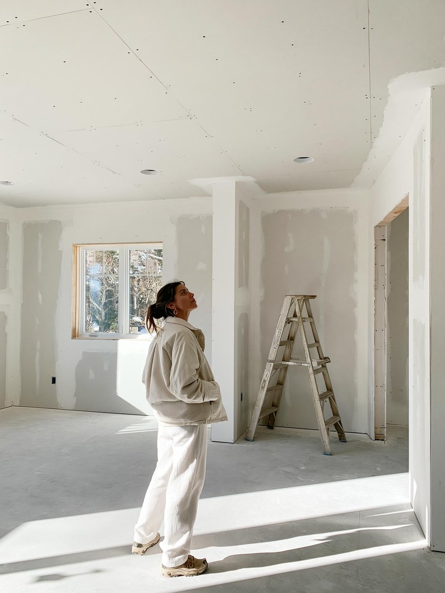 Homeowner standing in the middle of her new room addition with her hands in her pockets, turning to look up at the new drywall ceiling and along the new drywall walls of pale gray board finished in chalky white compound, a folded step ladder against one wall
