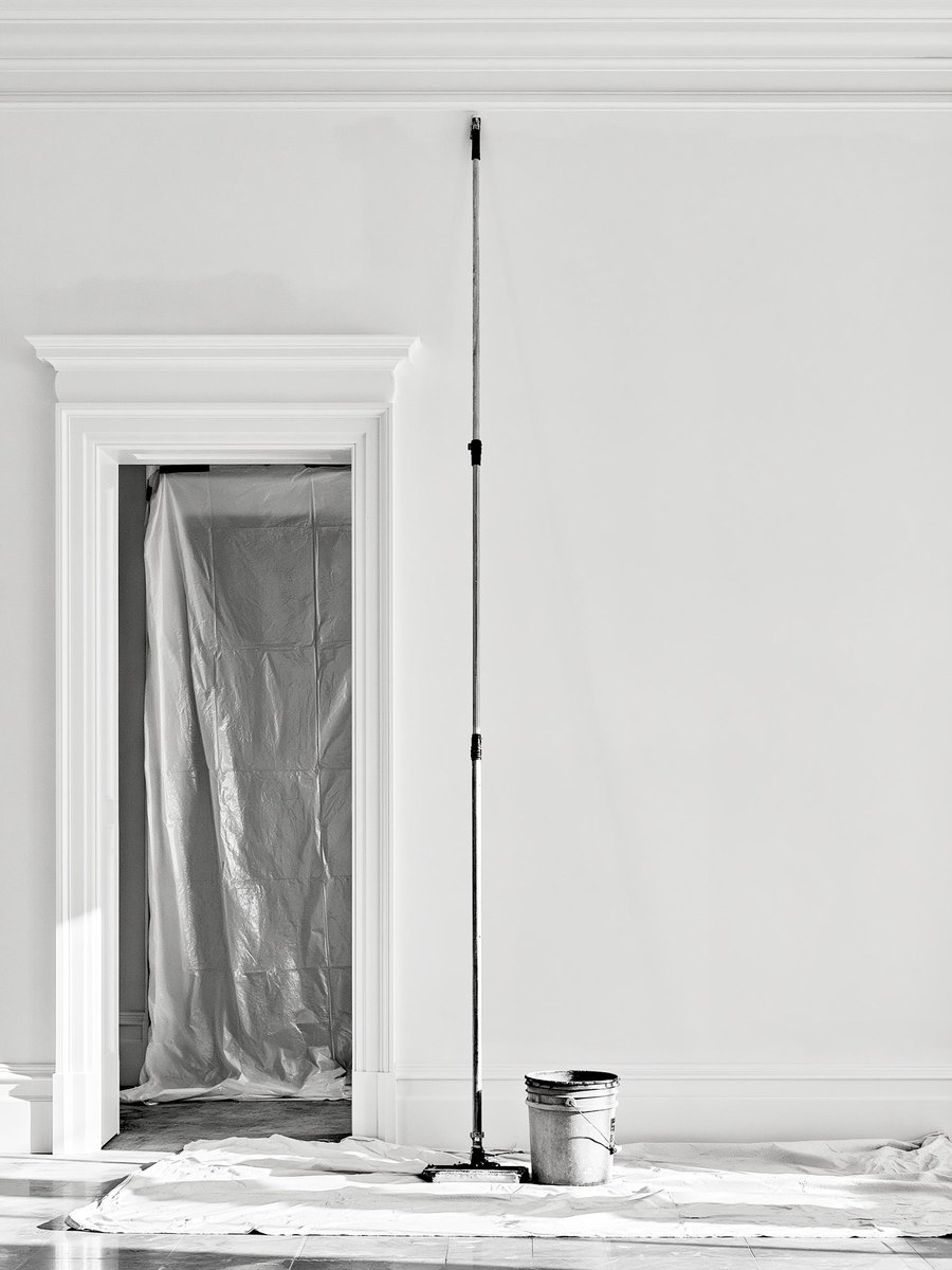 Sanding pole standing at rest against a freshly primed wall in a chalky warm off white, a lidded bucket beside it on a canvas drop cloth, the floor swept and a doorway masked with plastic sheeting behind