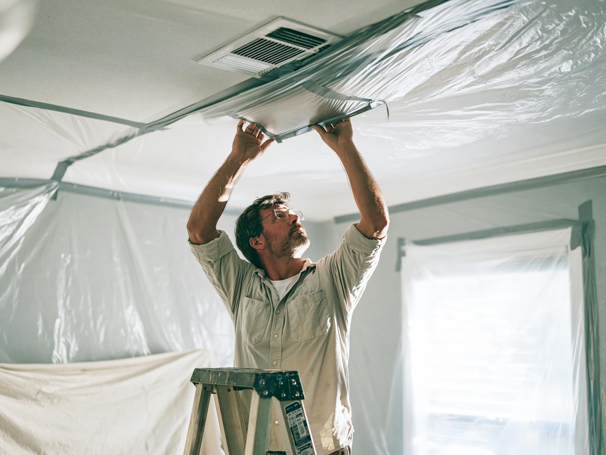 Man in a work shirt on a step stool in an emptied bedroom, pressing a square of plastic sheeting flat over a ceiling supply vent and smoothing the tape down, plastic already taped up the walls behind him and a canvas drop cloth over the carpet