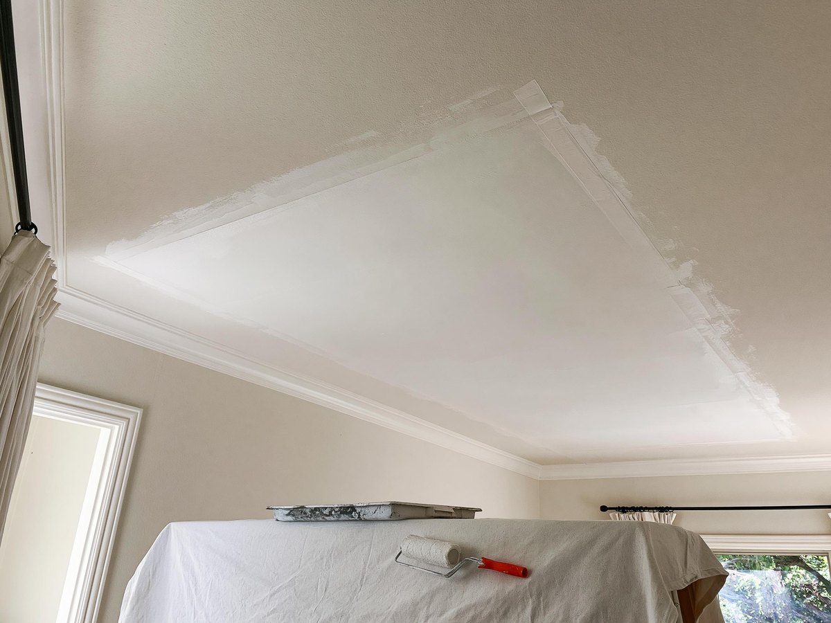 Rectangle of new ceiling board sealed and primed in chalky white against the surrounding warm off white bedroom ceiling, its taped edges feathered smooth into the ceiling around it, a paint roller at rest in its tray on a canvas drop cloth below