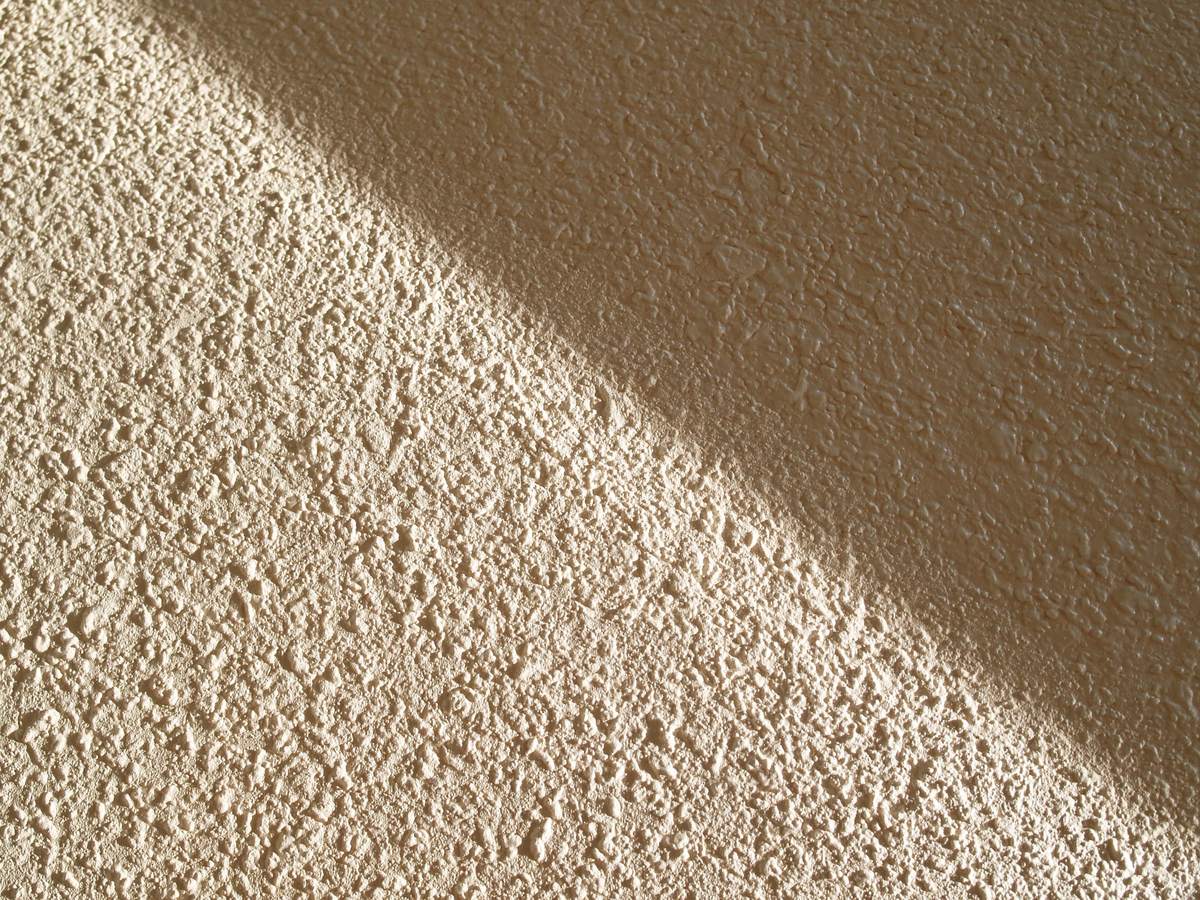 Close view along an orange peel textured bedroom wall painted a warm sand off white, where a finished repair is feathered out so far and so thin that the edge of it cannot be found, low daylight crossing the surface and the texture running unbroken through where the blend ends