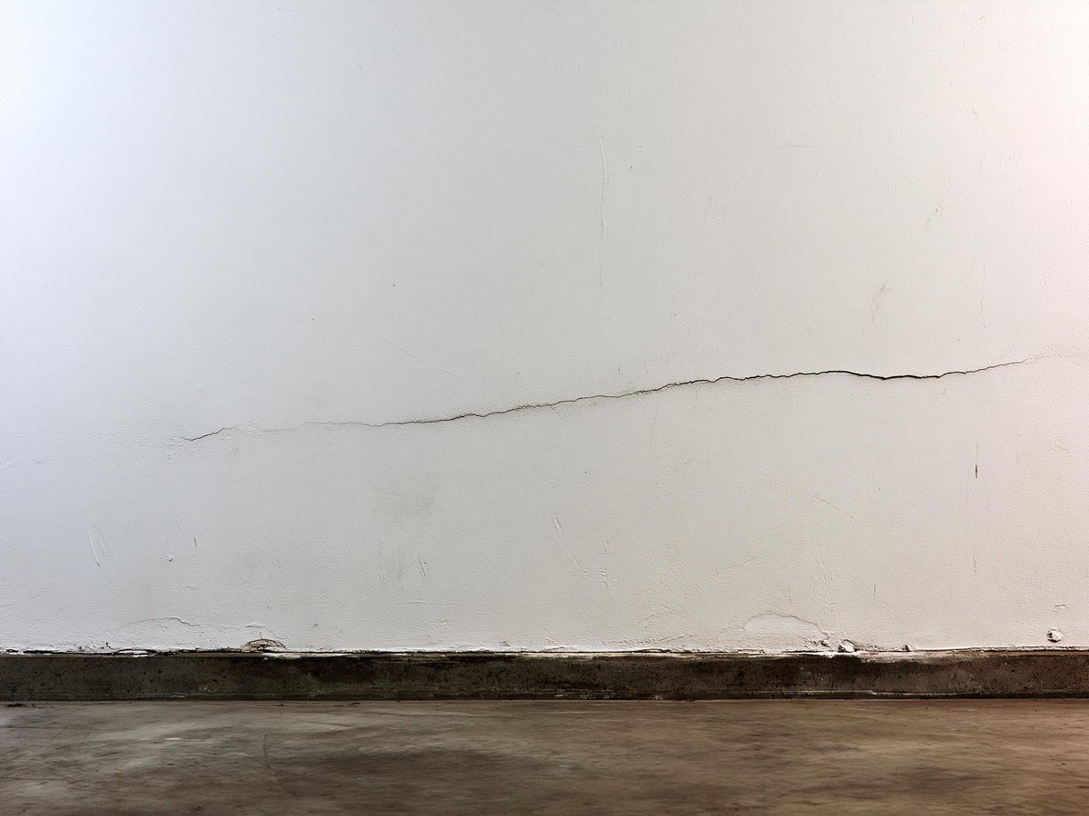 Close view along an interior garage drywall wall finished and painted a pale warm gray, scuffed and dented in a shallow horizontal line at the height of a car door, the sealed concrete floor meeting the wall at the bottom of the frame