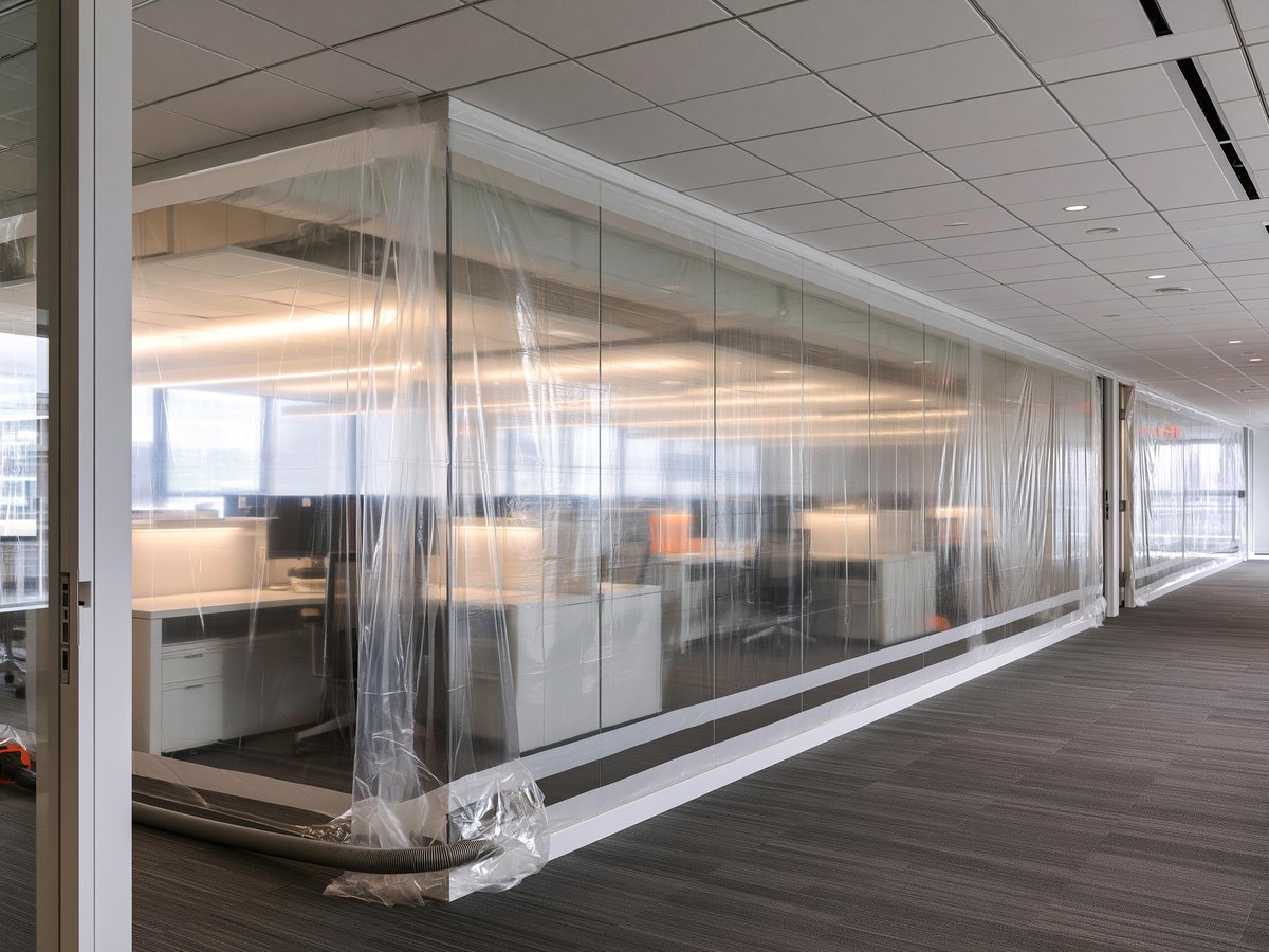 Clear plastic sheeting taped floor to ceiling across an office corridor, sealing the work area off from the working side, desks and task lighting glowing softly through it and a vacuum hose running along its base