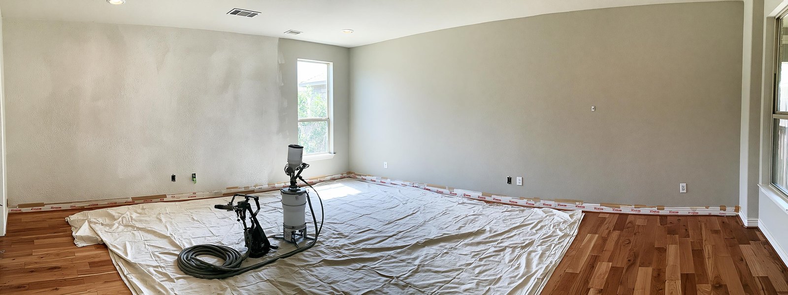 Living room masked off for texture work, paper and tape run along the baseboards and around the window trim, canvas drop cloths across the swept floor and a texture hopper gun standing upright at rest with its hose coiled beside it, one wall freshly sprayed in unpainted white orange peel texture