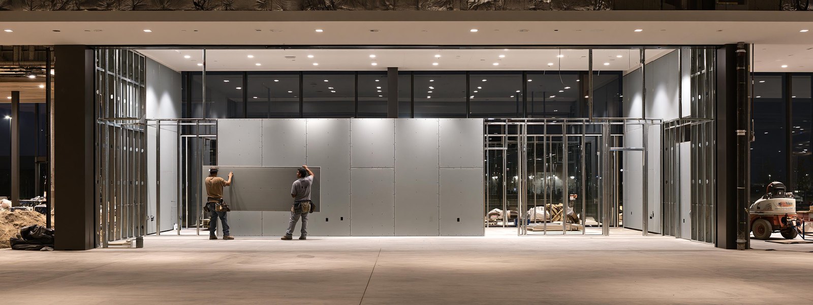 Two contractors in work shirts setting a full sheet of pale gray drywall board flat against the metal framing of a new partition wall inside an empty retail unit at night, the swept concrete sales floor running wide and open beside them and tall dark glazing behind