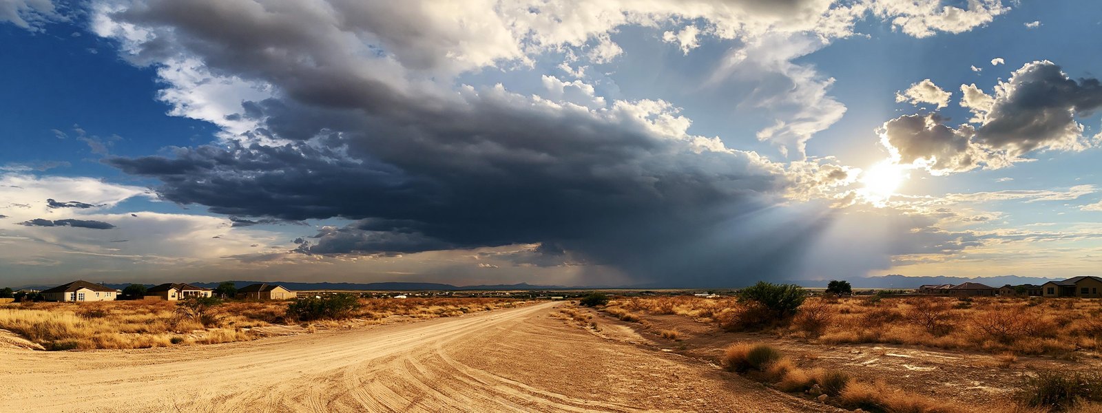 Open caliche and honey mesquite ranch ground where a new pale street runs out and stops, a clearing storm with breaking cloud and sun coming through filling the upper half of the frame, low new houses small and secondary along one edge