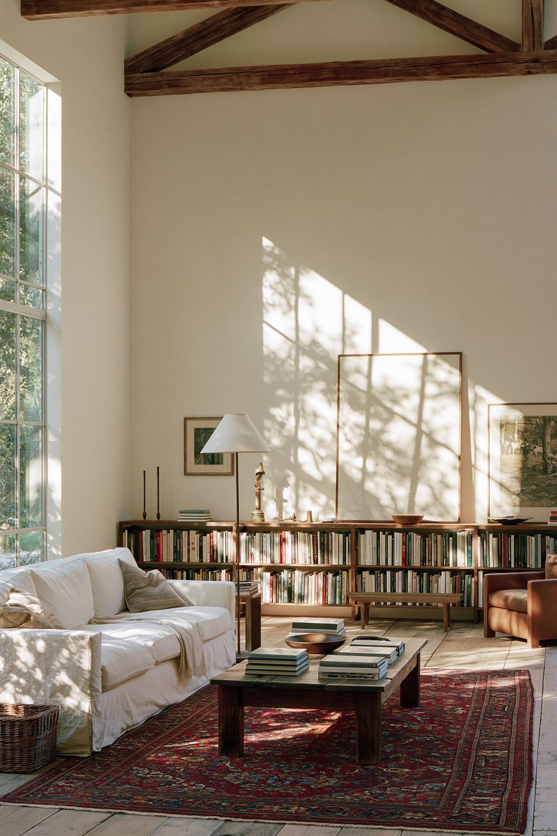 Long living room wall painted a warm off white, reading as one even surface, with a low bookshelf and a floor lamp against it and daylight from a tall window