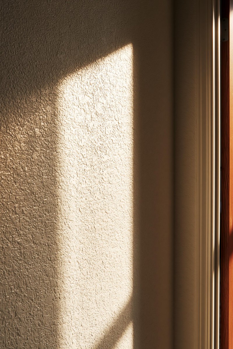 Knockdown textured wall painted warm off white, late sun skimming along the surface so the flattened plates of texture stand out, edge of a doorway at one side
