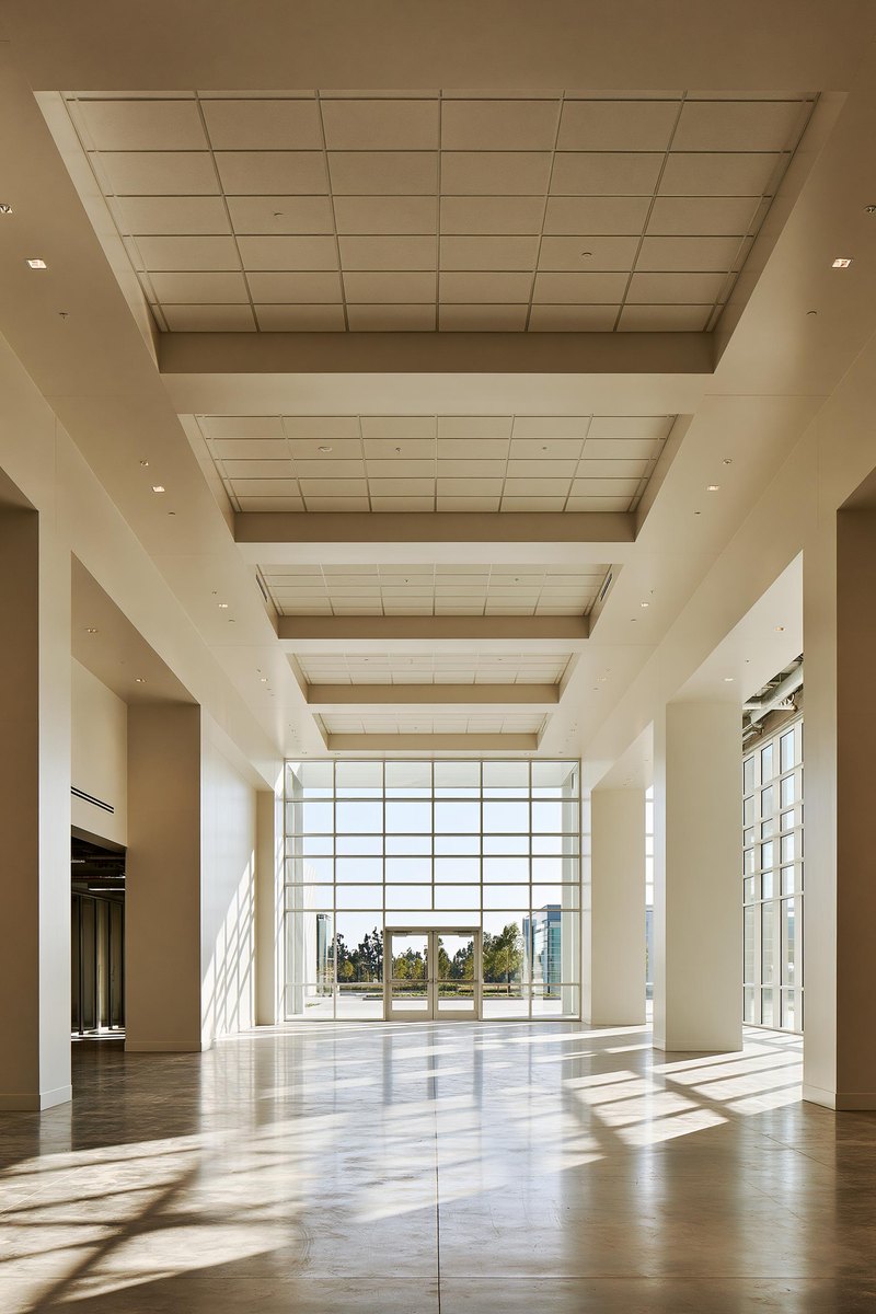 Finished commercial interior with warm off white walls running the length of the frame under a suspended ceiling grid, daylight from full height glazing at one end